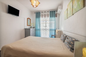 Ein Schlafzimmer mit einem Bett, einem Schminktisch, einem Spiegel, einem an der Wand befestigten Fernseher und einem Fenster mit Vorhängen.