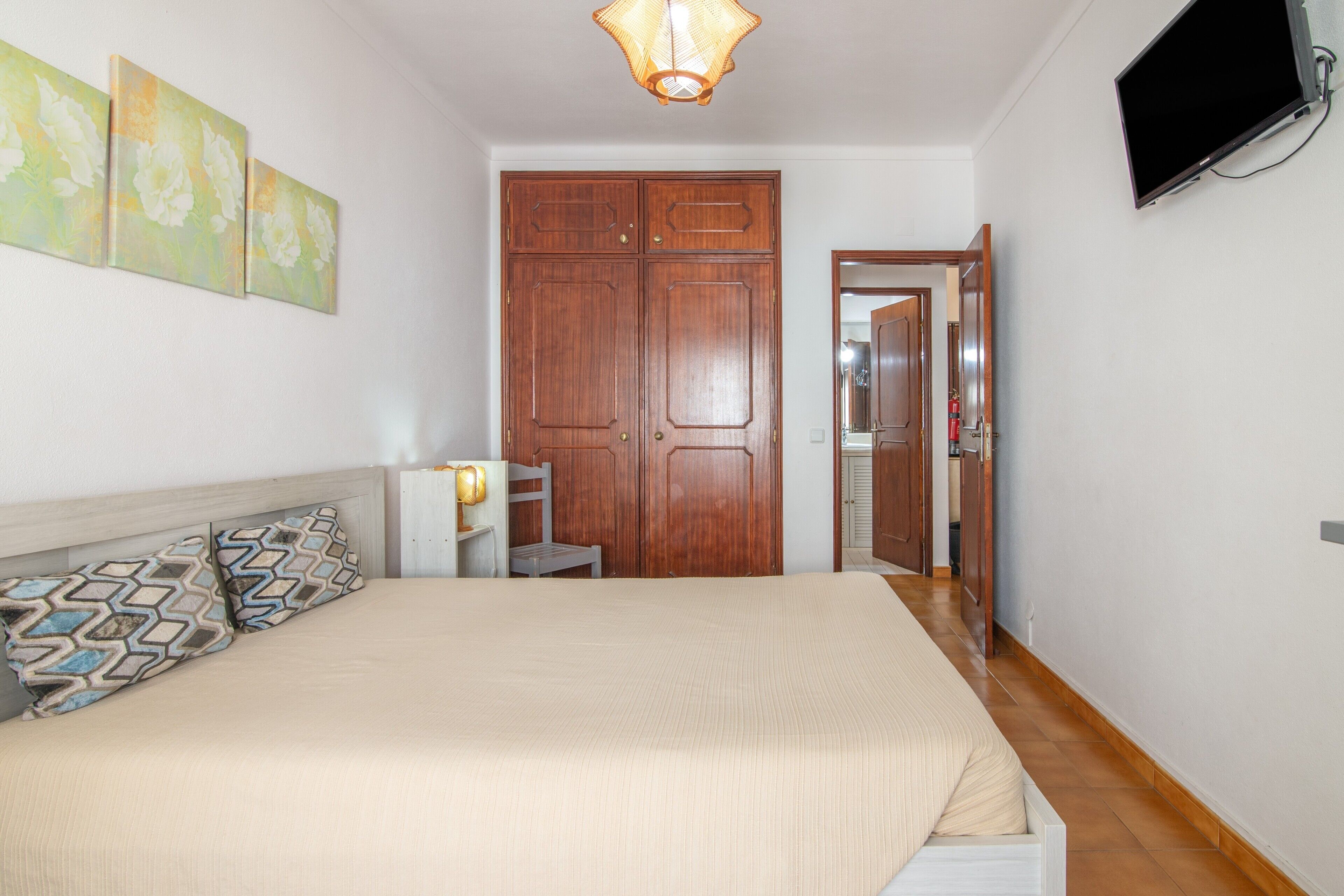A bedroom with a bed, a nightstand, a lamp, a wardrobe, and a television mounted on the wall.