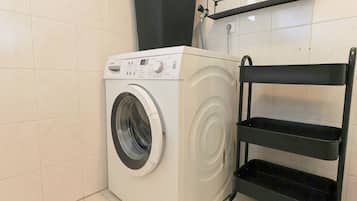 A front-loading washing machine in a bathroom with tiled walls and a black shelf unit.