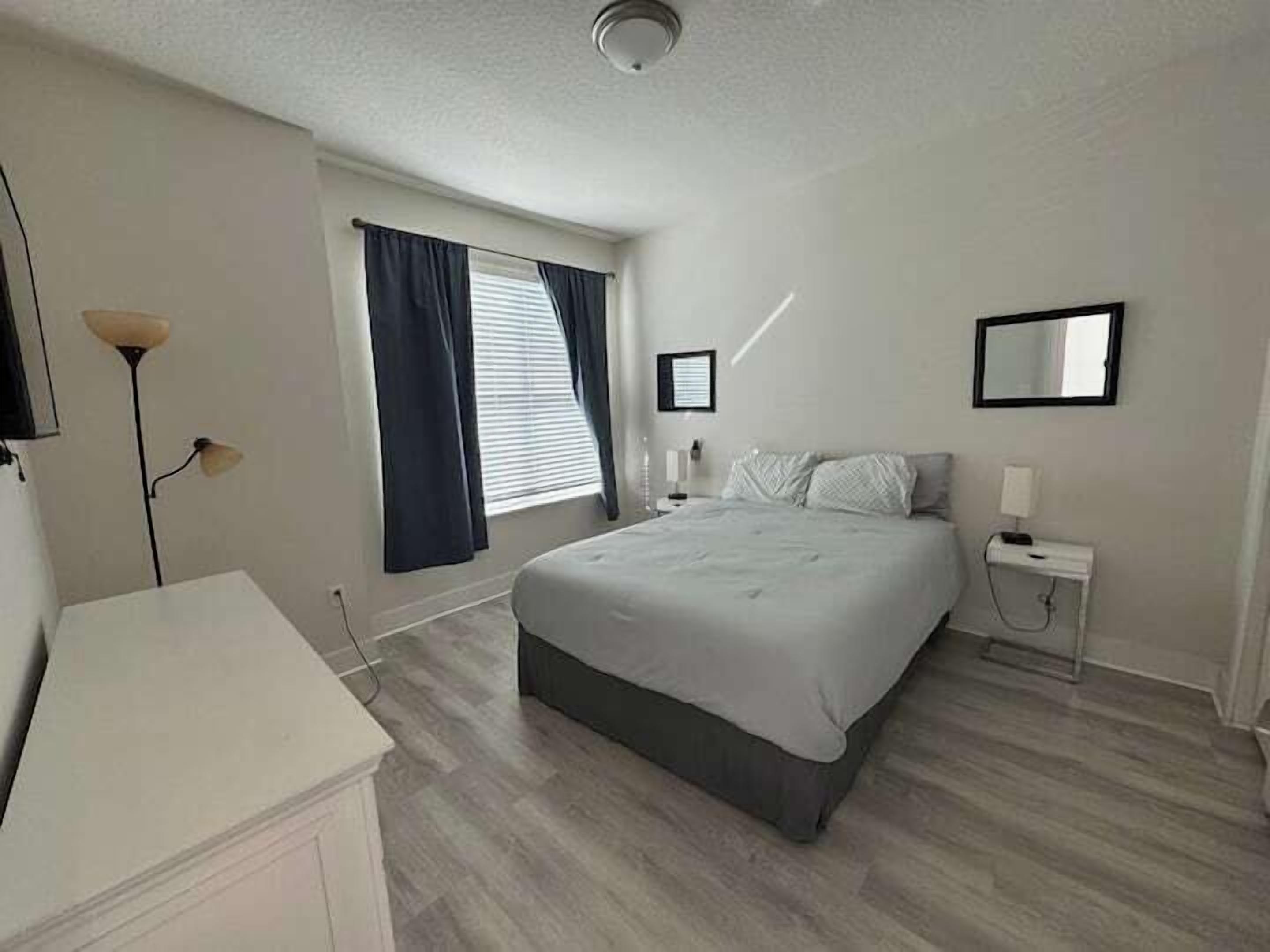 A bedroom with a bed, bedside table, lamp, window with curtains, and a mirror on the wall.