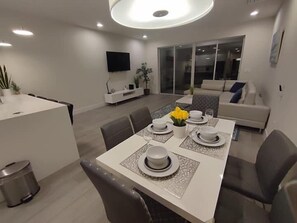 A modern dining area with a long table set for four, a sofa in the background, and a TV mounted on the wall. - Modern Townhome with Private Splash Pool, Near Disney (Clermont)