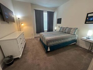 Une chambre à coucher avec un lit, une commode, un téléviseur et une fenêtre avec des rideaux.