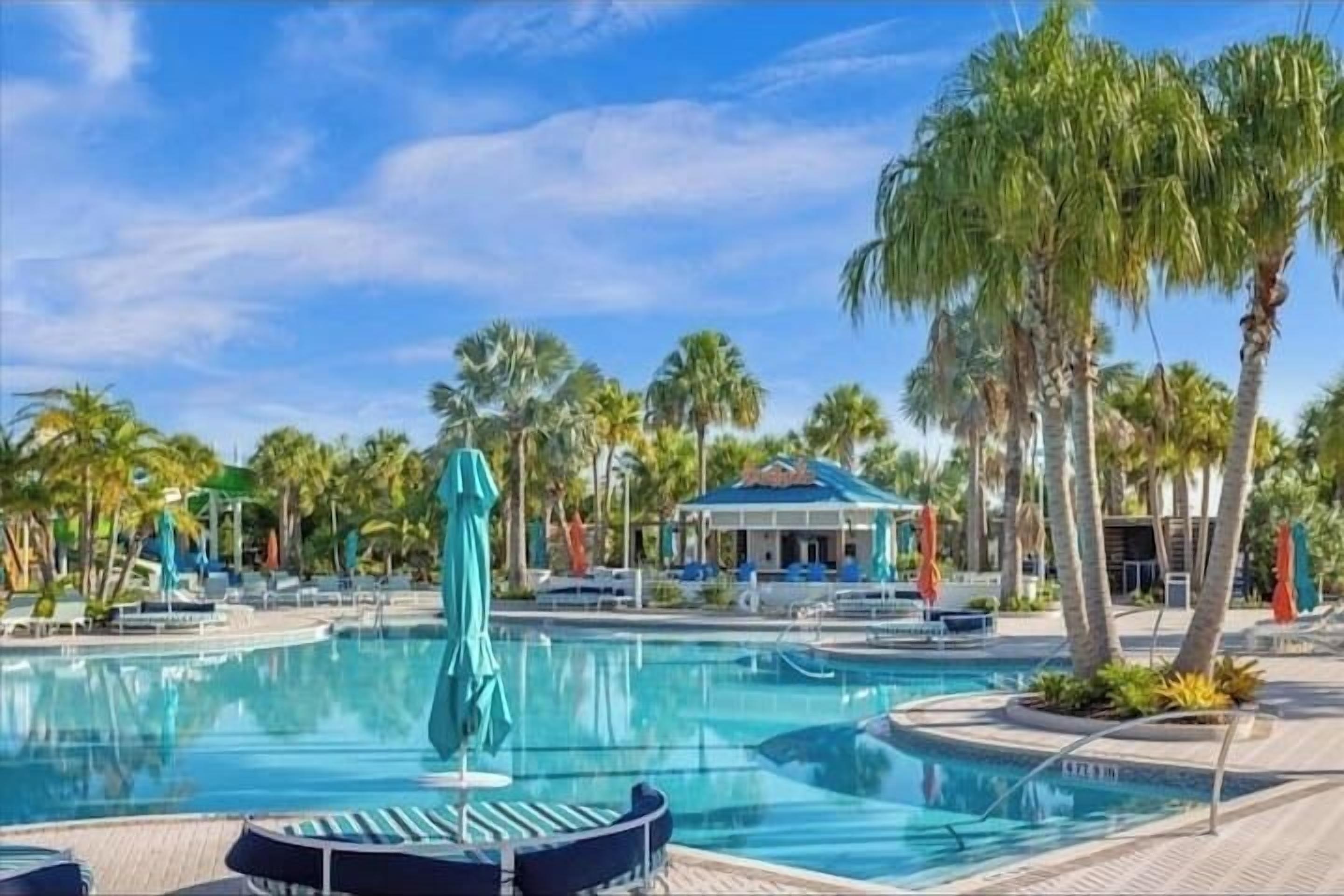 A swimming pool with lounge chairs and umbrellas, surrounded by palm trees and a building with a blue roof.