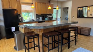 Uma cozinha moderna com armários de madeira, uma geladeira preta, bancada de granito, luminárias pendentes e um espaço de bar com banquetas.