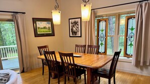 Uma sala de jantar com mesa e cadeiras de madeira, quadros nas paredes e vista para o verde lá fora.