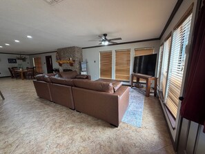 A spacious living room with a large brown sectional sofa, a stone fireplace, a ceiling fan, and a dining area with a wooden table and chairs. - Knot On Call- Peaceful 3-bedroom on Lake Texoma (Kingston)