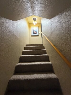 Una escalera alfombrada con barandilla que lleva a una plataforma con un cuadro enmarco y una lámpara.