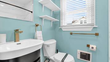 Une salle de bain avec un lavabo blanc, des toilettes et une fenêtre avec des stores. On y trouve des étagères blanches et un porte-serviettes.