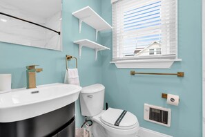 A bathroom with a white sink, toilet, and window with blinds. There are white shelves and a towel rack. - Prof Managed & Cleaned | Half Block to Beach (Seaside Heights)