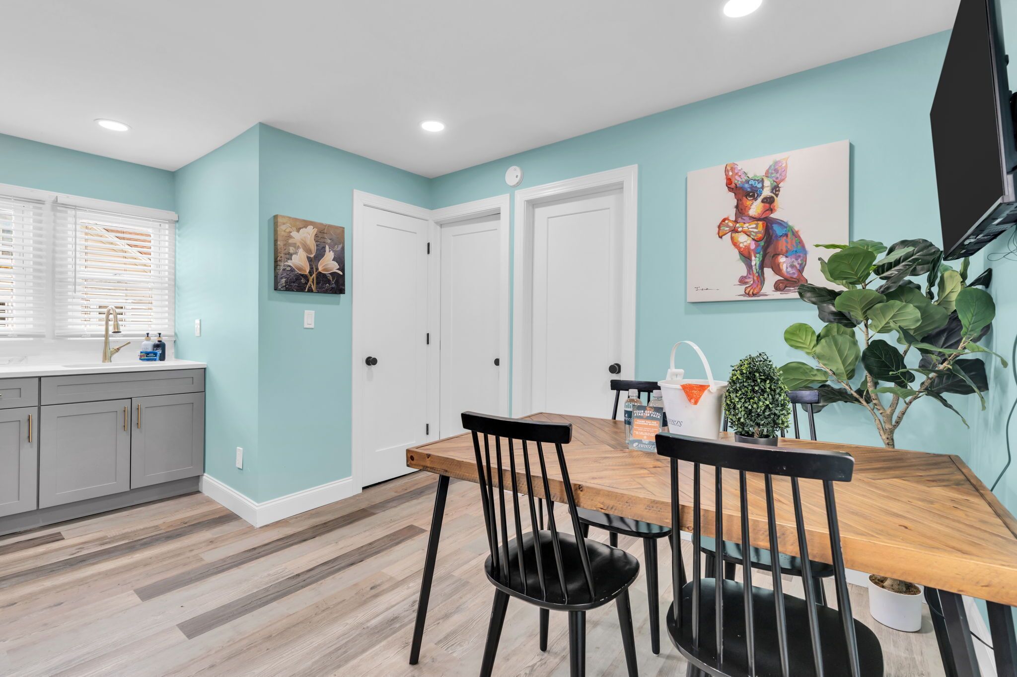 Un coin repas avec une table en bois et des chaises noires, une cuisine avec des meubles gris et un mur de couleur turquoise orné d’œuvres d’art.