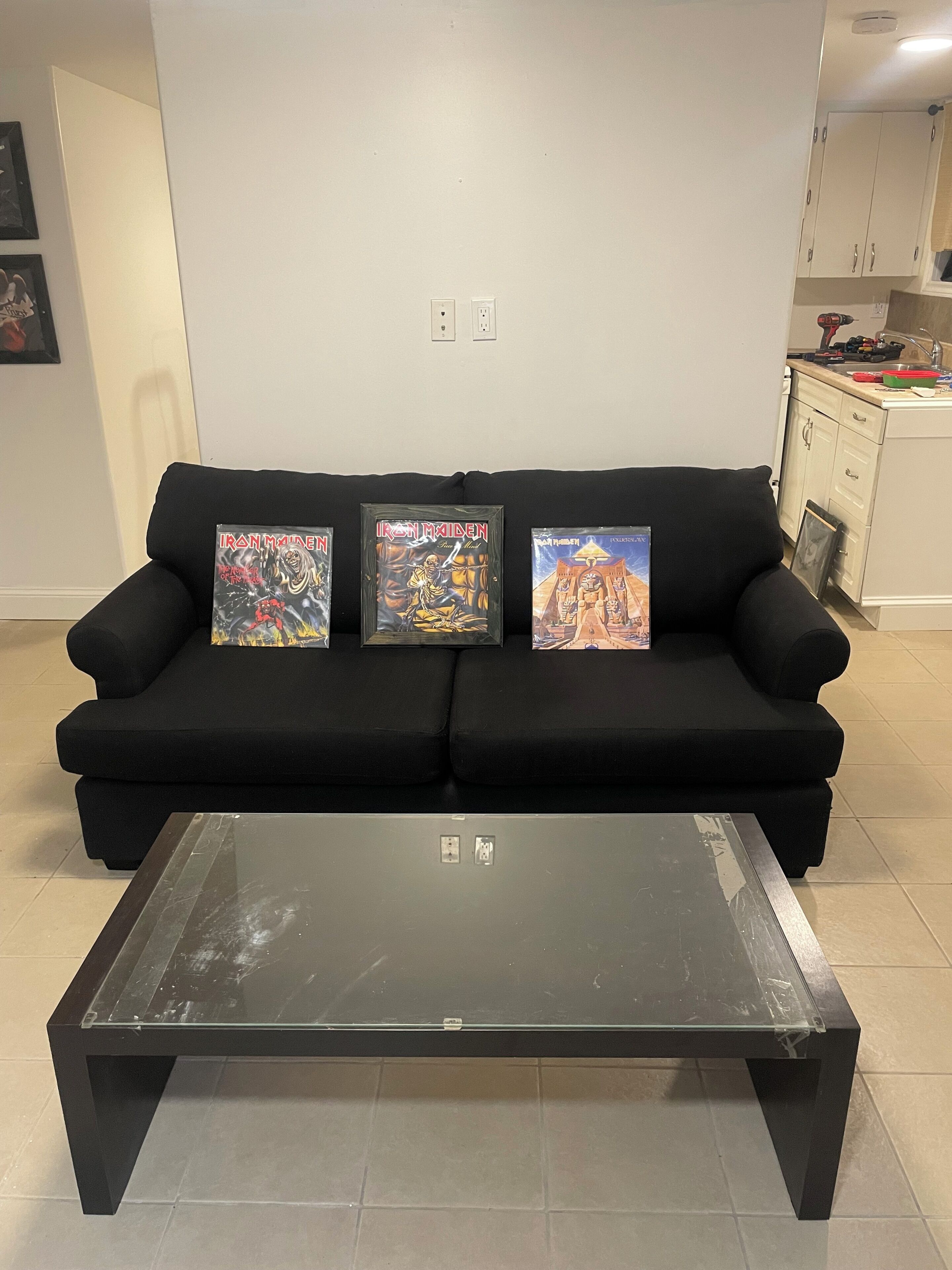 A black sofa with three framed Iron Maiden albums against a plain wall, a glass-top coffee table in front, and a kitchen area to the right.
