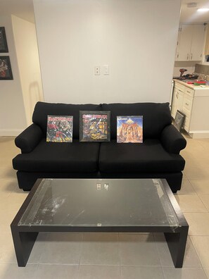 Un sofá negro con tres álbumes enmarcados de Iron Maiden sobre una pared lisa, una mesa de centro con superficie de vidrio al frente, y una zona de cocina a la derecha.