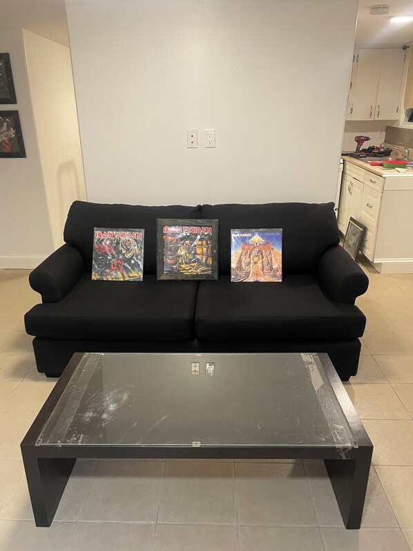 A black sofa with three framed Iron Maiden albums against a plain wall, a glass-top coffee table in front, and a kitchen area to the right.