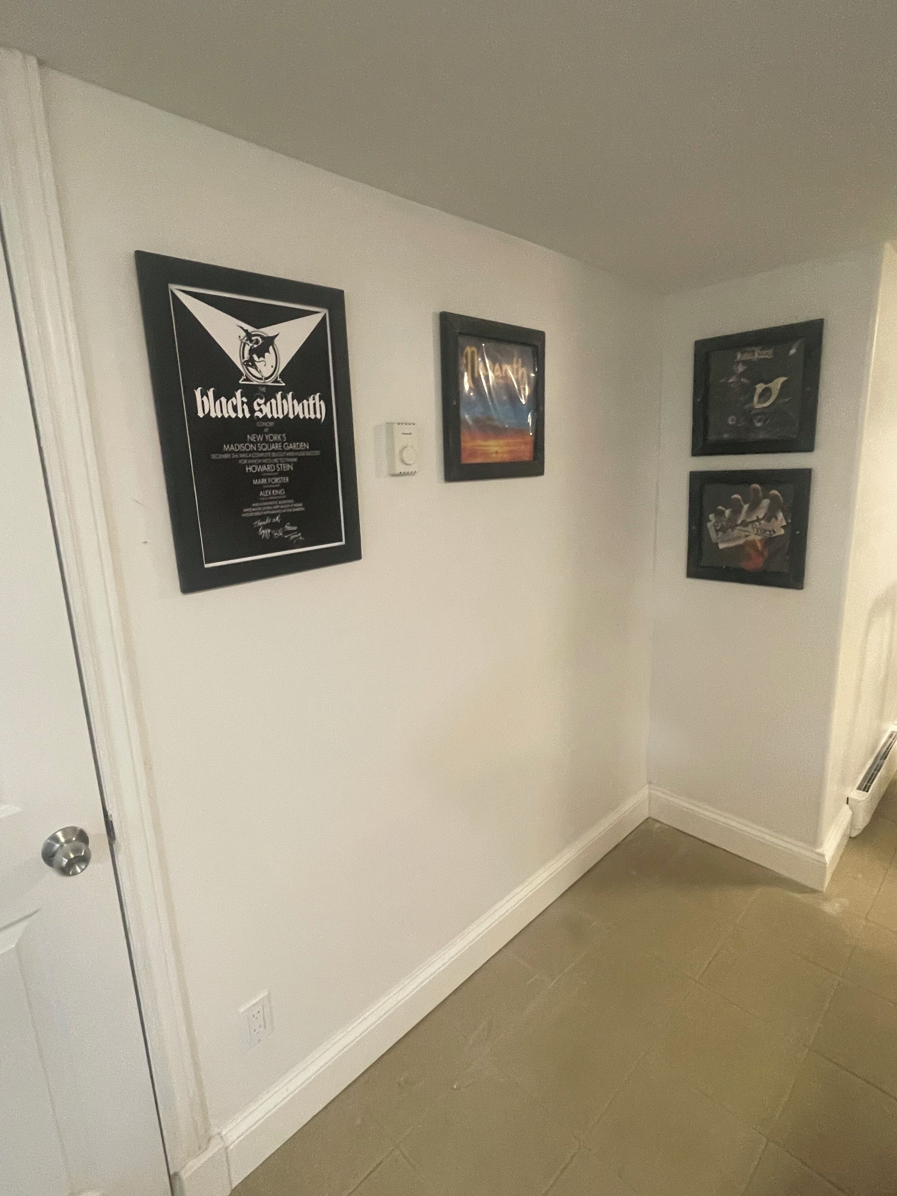 Un couloir aux murs blancs et au sol carrelé. Trois objets encadrés sont accrochés au mur : un panneau noir avec du texte blanc, une image encadrée représentant un coucher de soleil et une image encadrée représentant un oiseau.