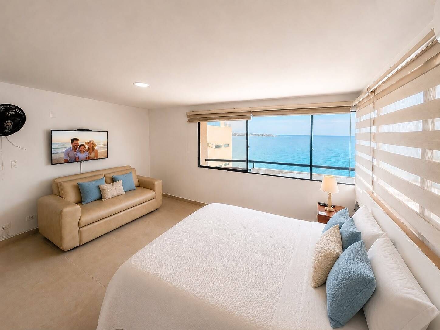 A bedroom with a bed, a sofa, a TV, and a window with a view of the sea.