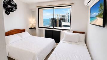 Een hotelkamer met twee bedden, een raam met uitzicht op zee en gebouwen, een luchtverkoeler aan de muur, en een plafondventilator.