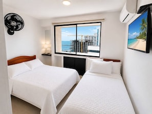 Een hotelkamer met twee bedden, een raam met uitzicht op zee en gebouwen, een luchtverkoeler aan de muur, en een plafondventilator.