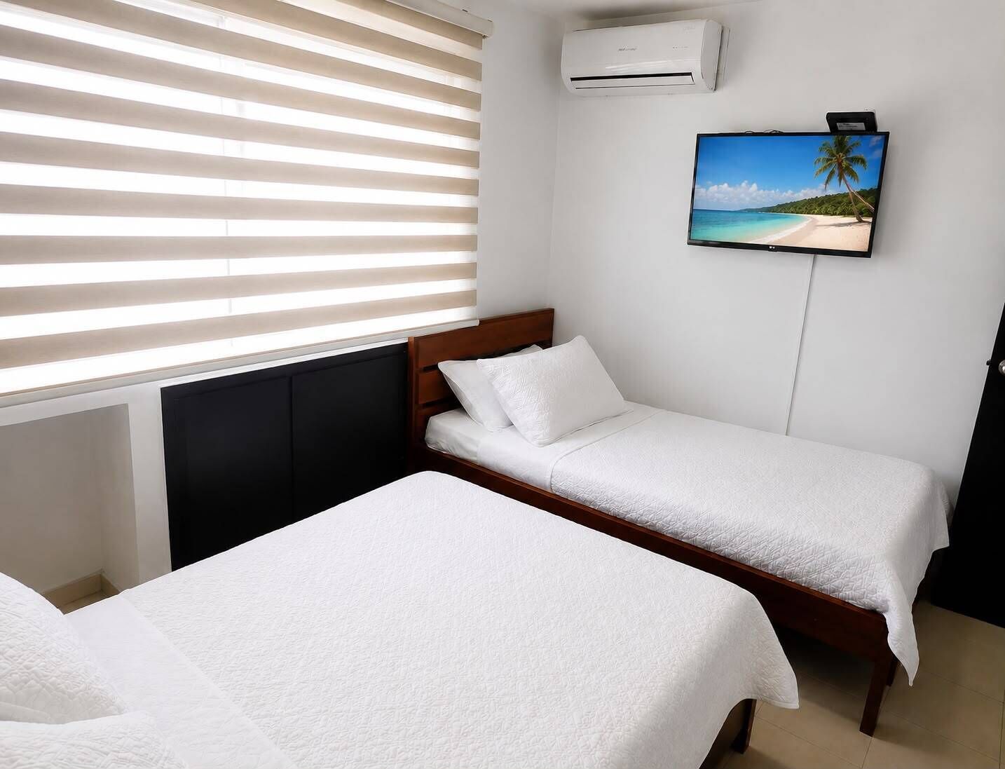 A room with two single beds, a wall-mounted air conditioner, and a TV displaying a beach scene.