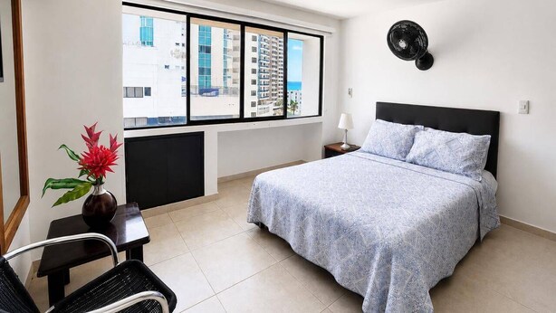 Quarto de hotel com cama, cadeira, mesinha, um vaso de flores e janela com vista para a cidade.