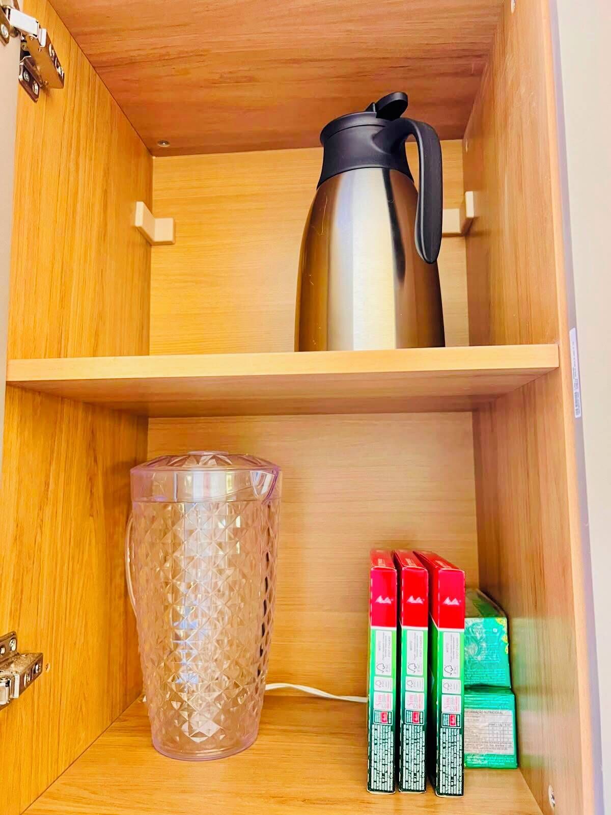 Uma prateleira de madeira com uma cafeteira de aço inoxidável, um recipiente de vidro transparente e várias caixas verdes e vermelhas.