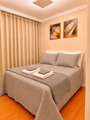Una cama bien hecha con una colcha gris y almohadas, un par de toallas dobladas sobre la cama y dos piezas decorativas de plumas en la pared encima.