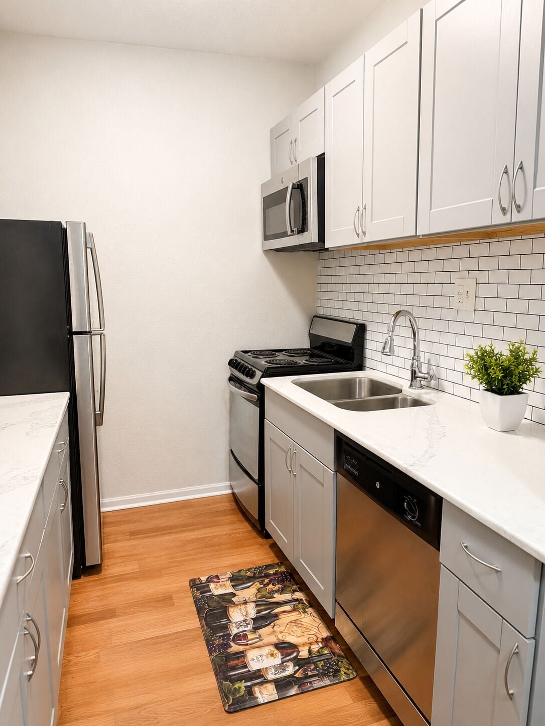 A compact kitchen with stainless steel appliances, white cabinets, a tiled backsplash, and a wooden floor.