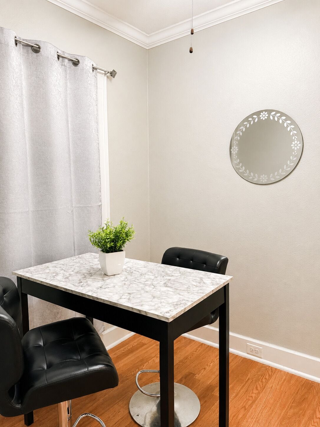 A small, modern room with a marble table, two black chairs, a potted plant, a round mirror, and a curtain.