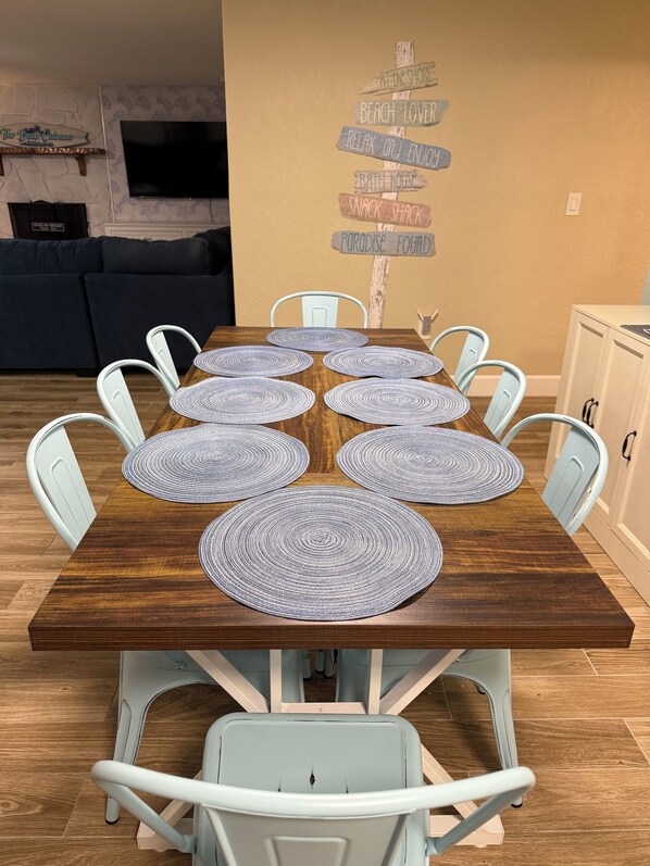 A dining table with placemats, surrounded by white chairs, in a room with a wall-mounted TV and a sign with various phrases.