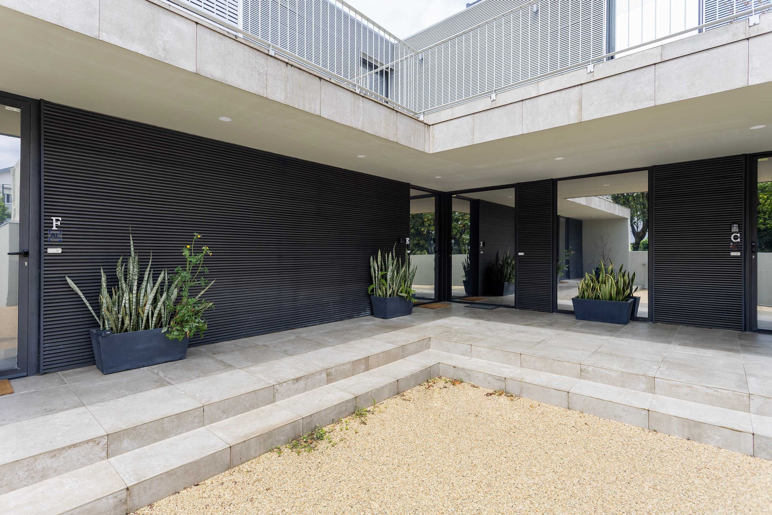 Un bâtiment moderne doté d’un espace extérieur couvert comprenant une terrasse en gravier, des plantes en pot et un escalier menant à un niveau supérieur.