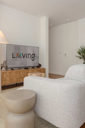 Ein modernes Wohnzimmer mit Flachbildfernseher auf einer Holzkonsole, einem grauen Polstersofa, einem runden Couchtisch und einer Zimmerpflanze.