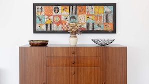 Een houten kast met daarboven een lijst met een abstract schilderij, een vaas met gedroogde bloemen en twee kommen op de bovenkant.