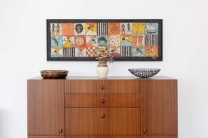 Ein Holzschrank mit einem dahinter hängenden, gerahmten abstrakten Gemälde, einer Vase mit Trockenblumen und zwei Schalen darauf.