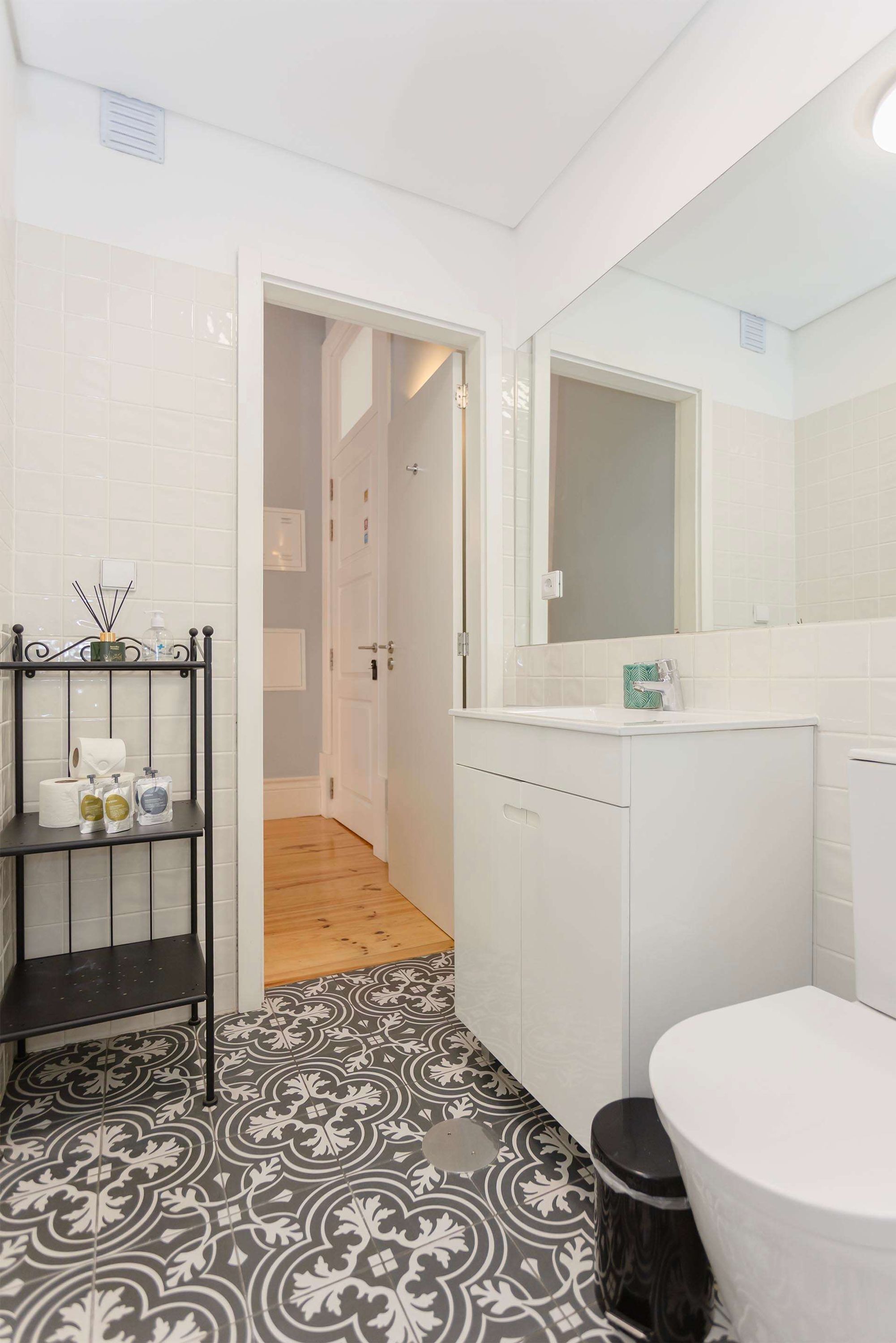 Une salle de bain avec des toilettes blanches, un lavabo avec un miroir, une étagère avec des articles de toilette et des carreaux de sol à motifs.