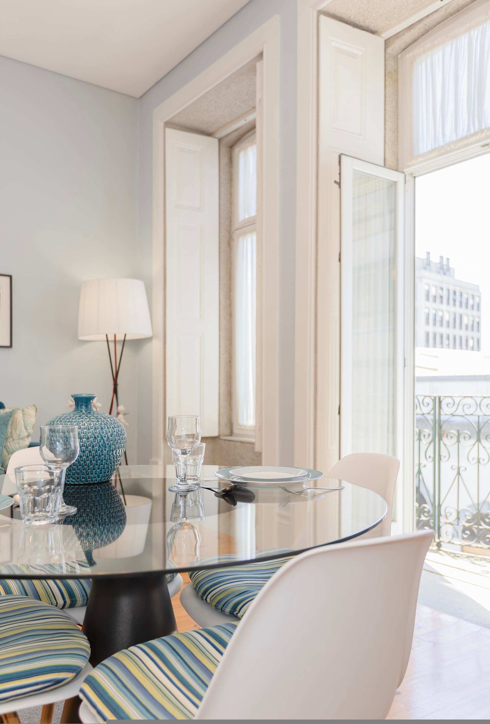 A modern dining area with a round glass table, striped cushions, and a view of the city through large windows.