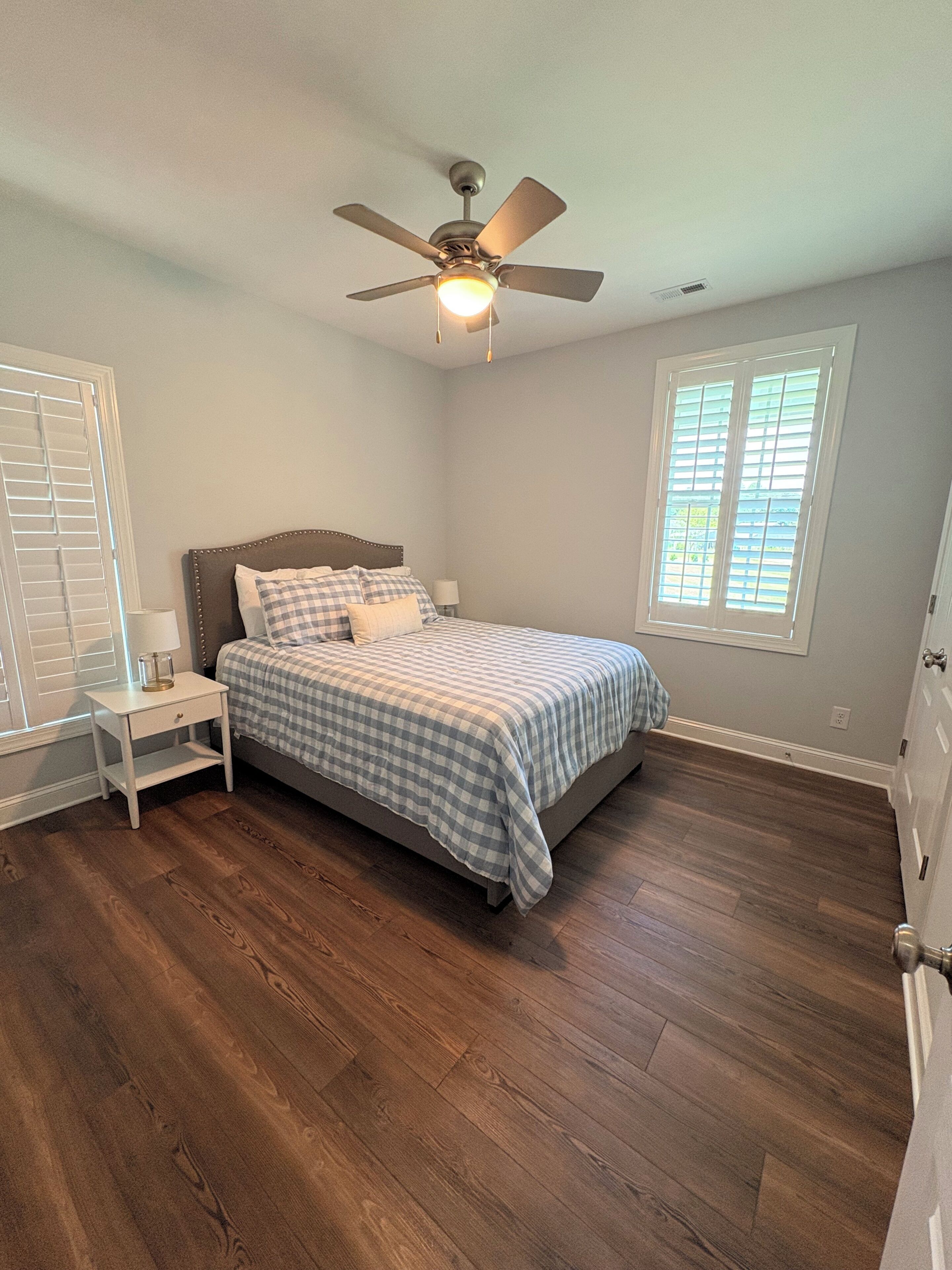 A bedroom with a bed, nightstand, ceiling fan, and a window with blinds.