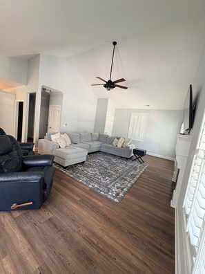 Ein Wohnzimmer mit einem Ecksofa, einem schwarzen Ledersessel, einem gemusterten Teppich und einem Deckenventilator.
