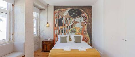 Un dormitorio con cama, una banca, una mesita y un mural en la pared.