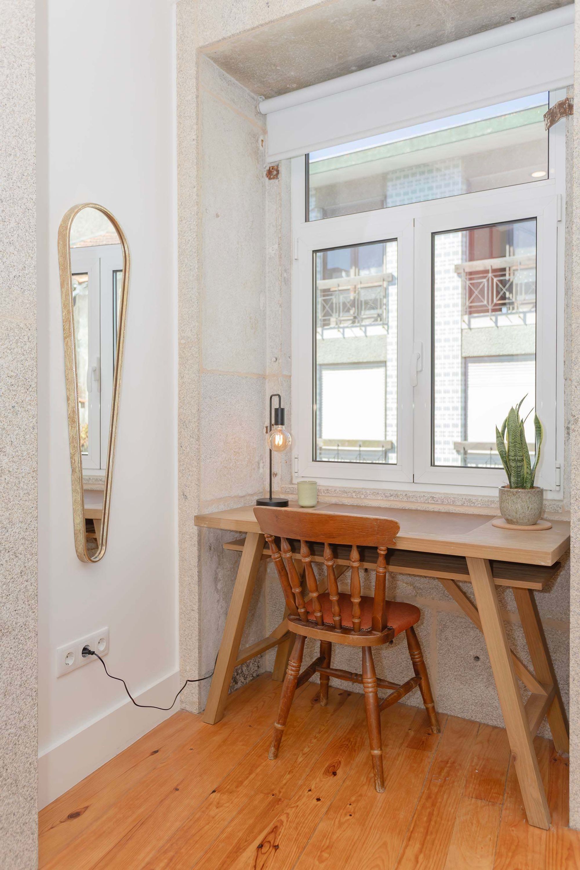 Een hoekkamer met een houten bureau, een stoel, een spiegel, een raam en een plant.