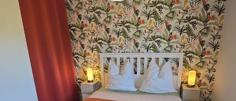 A bed with a white headboard, a red bedspread, and two white towels. There are two bedside lamps with lit candles. The wall behind the bed has a tropical floral wallpaper.