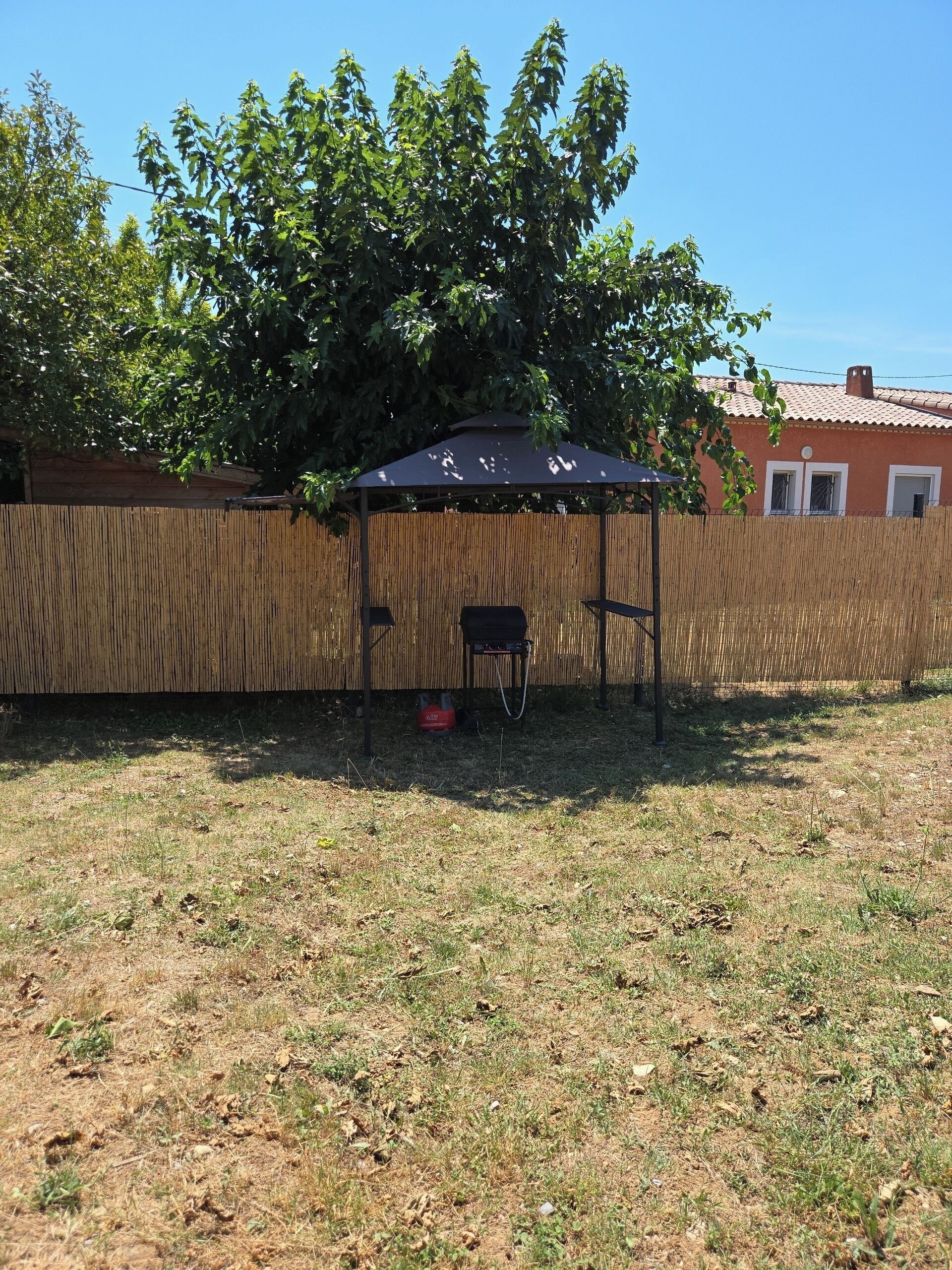Een overdekt terras met een bamboehek, een rode gasfles en een huis op de achtergrond.