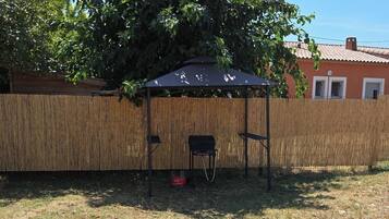 Een overdekt terras met een bamboehek, een rode gasfles en een huis op de achtergrond.