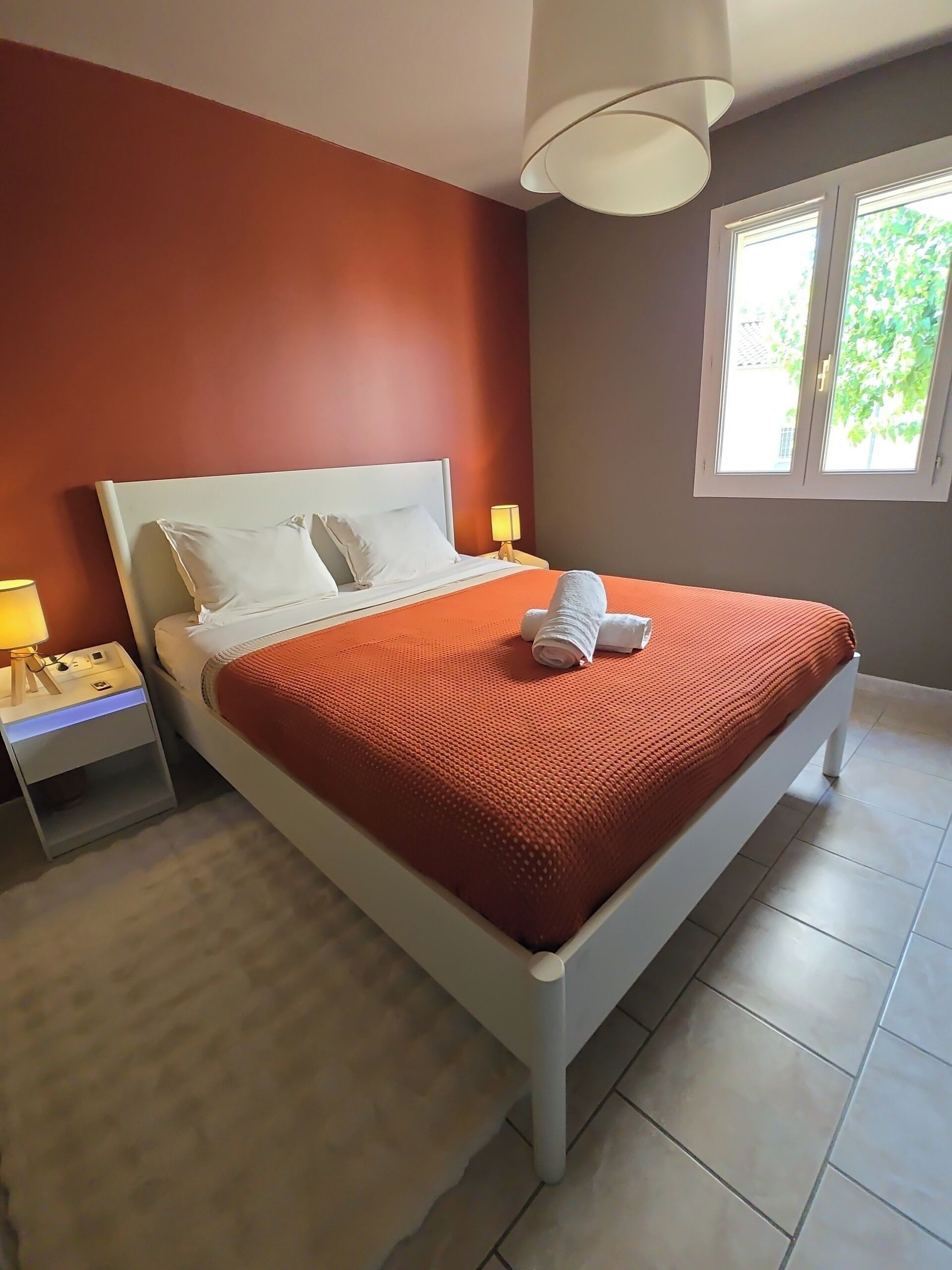 A bed with a white headboard, a red blanket, and two pillows. There is a nightstand with a lamp and a power outlet. A window with a view of greenery is visible.