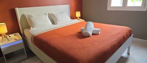 A bed with a white headboard, a red blanket, and two pillows. There is a nightstand with a lamp and a power outlet. A window with a view of greenery is visible.