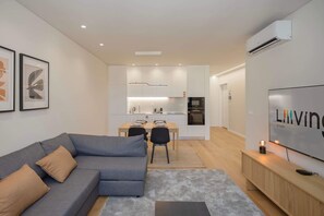 A modern living room with a grey sofa, a flat-screen TV, and a dining area with a kitchen in the background. - Liiiving in Matosinhos - Mercatus Apartment C (Matosinhos)