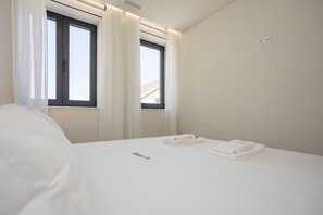 A hotel room with a bed, two windows with curtains, a wall socket, and a folded towel on the bed. - Liiiving in Matosinhos - Mercatus Apartment C (Matosinhos)