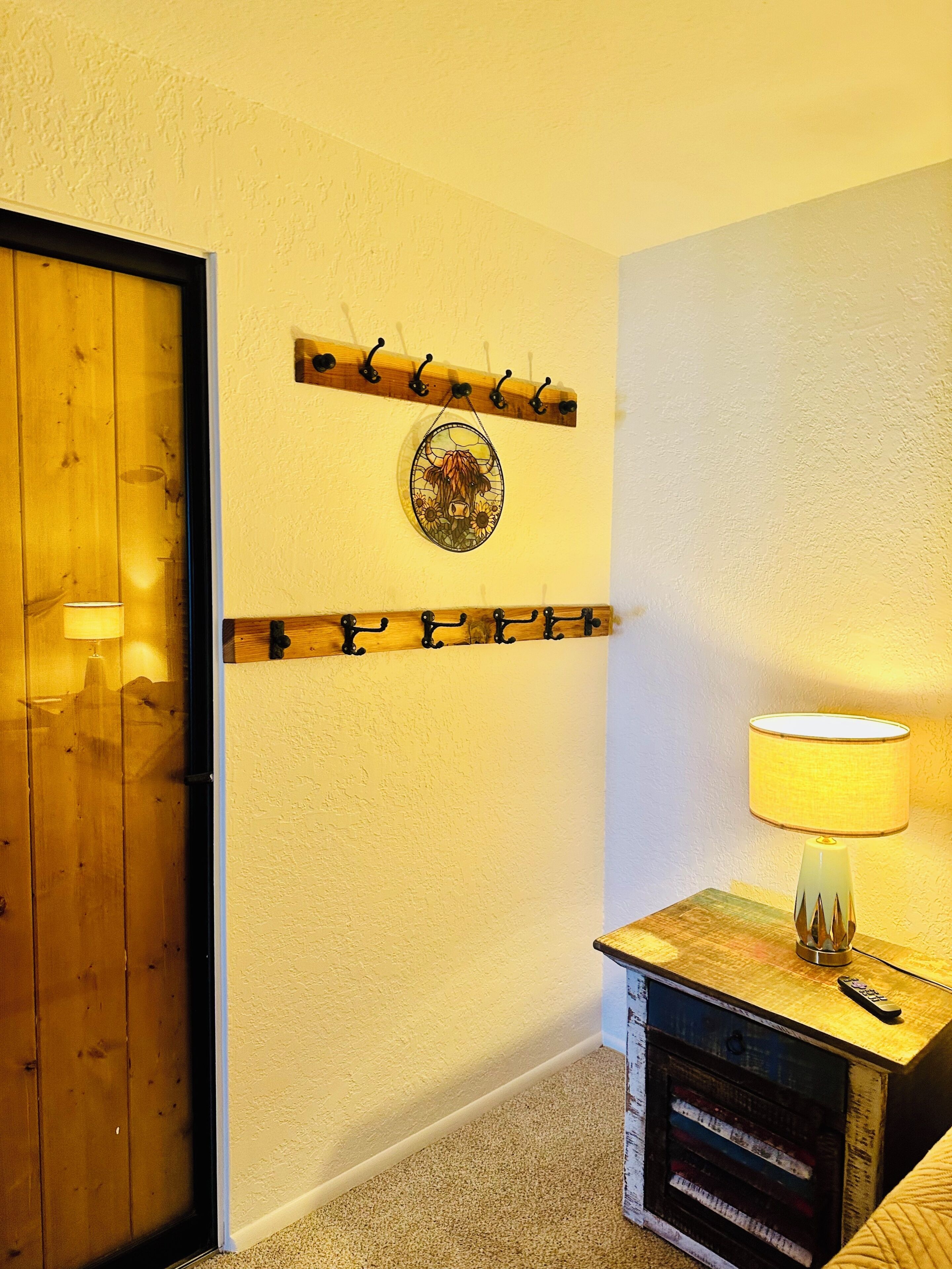 A room with a wooden door, a wall-mounted coat rack, a decorative plate, a lamp, and a small cabinet with drawers.