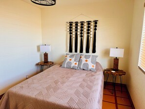 Un dormitorio con una cama, dos mesitas de noche, un adorno de macramé en la pared y una ventana con persianas.
