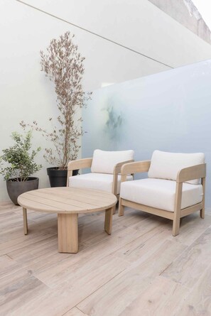 Ein Holztisch und zwei weiße Sitzgelegenheiten mit Kissen in einer modernen Außenanlage, ergänzt durch Pflanzgefäße.