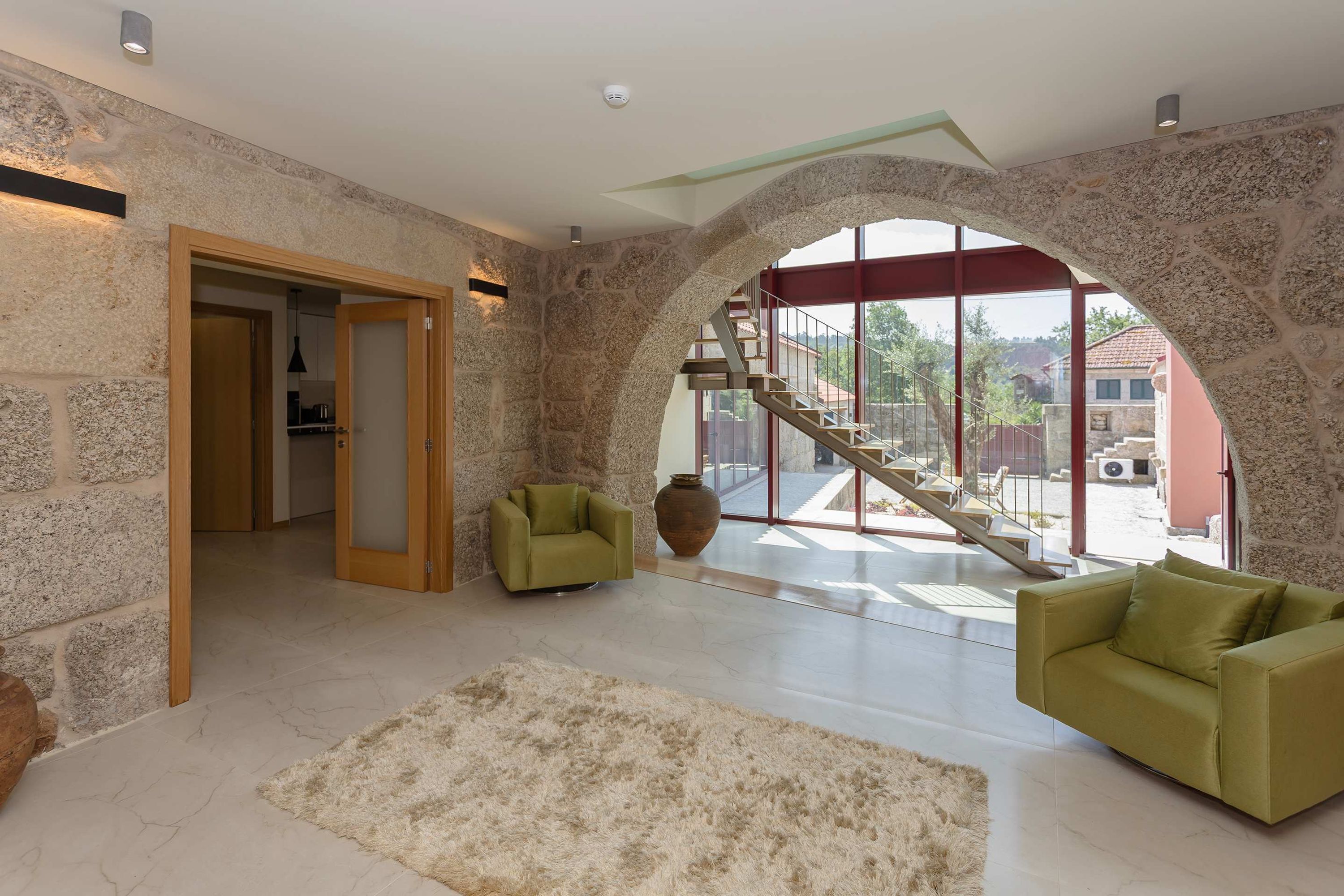 Amplia sala de estar con pared de piedra, puerta de madera, un sillón verde, alfombra beige y vista a una escalera que conduce al exterior.