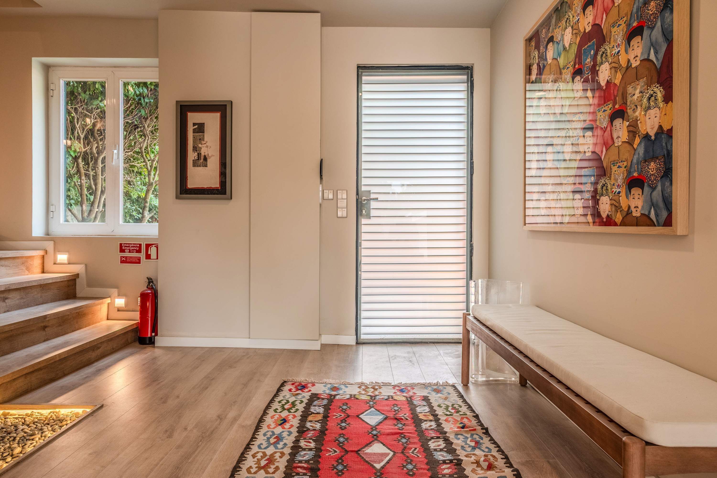 A modern interior with a staircase, a fire extinguisher, a patterned rug, a bench, and a door with blinds.